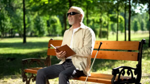 Happy blind pensioner reading funny story in braille book, enjoying rest in park